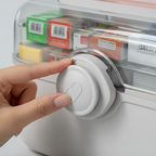 Hand pressing a button on a white electronic device with a clear container holding labeled boxes in the background.